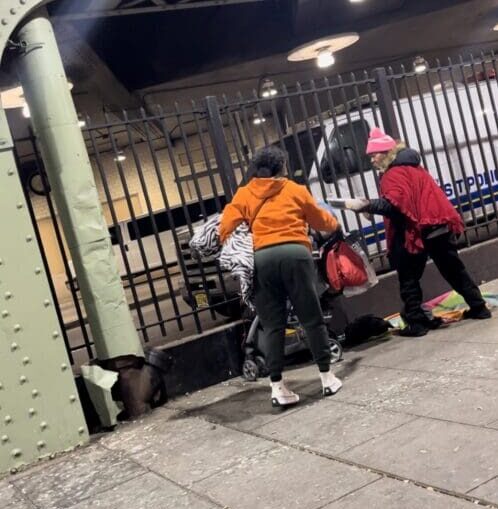 Two people are walking on a sidewalk near a metal fence.