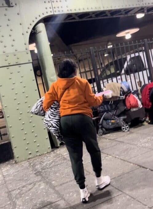 A woman in orange jacket walking on sidewalk next to metal fence.