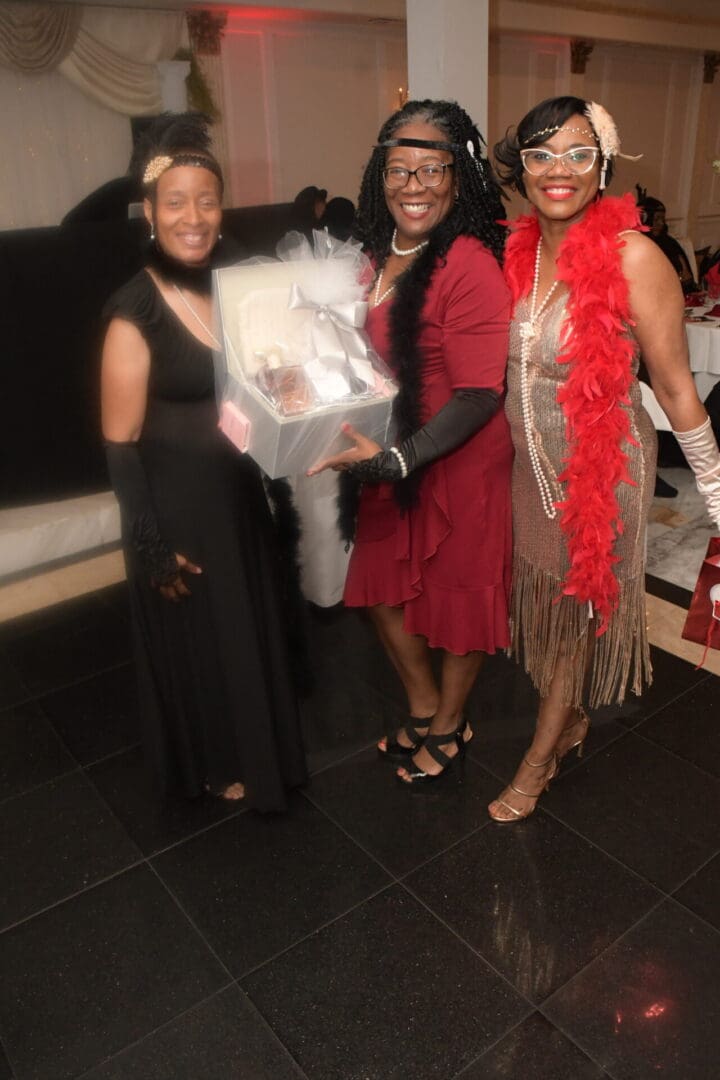 Three women in black and red dresses holding a white gift.